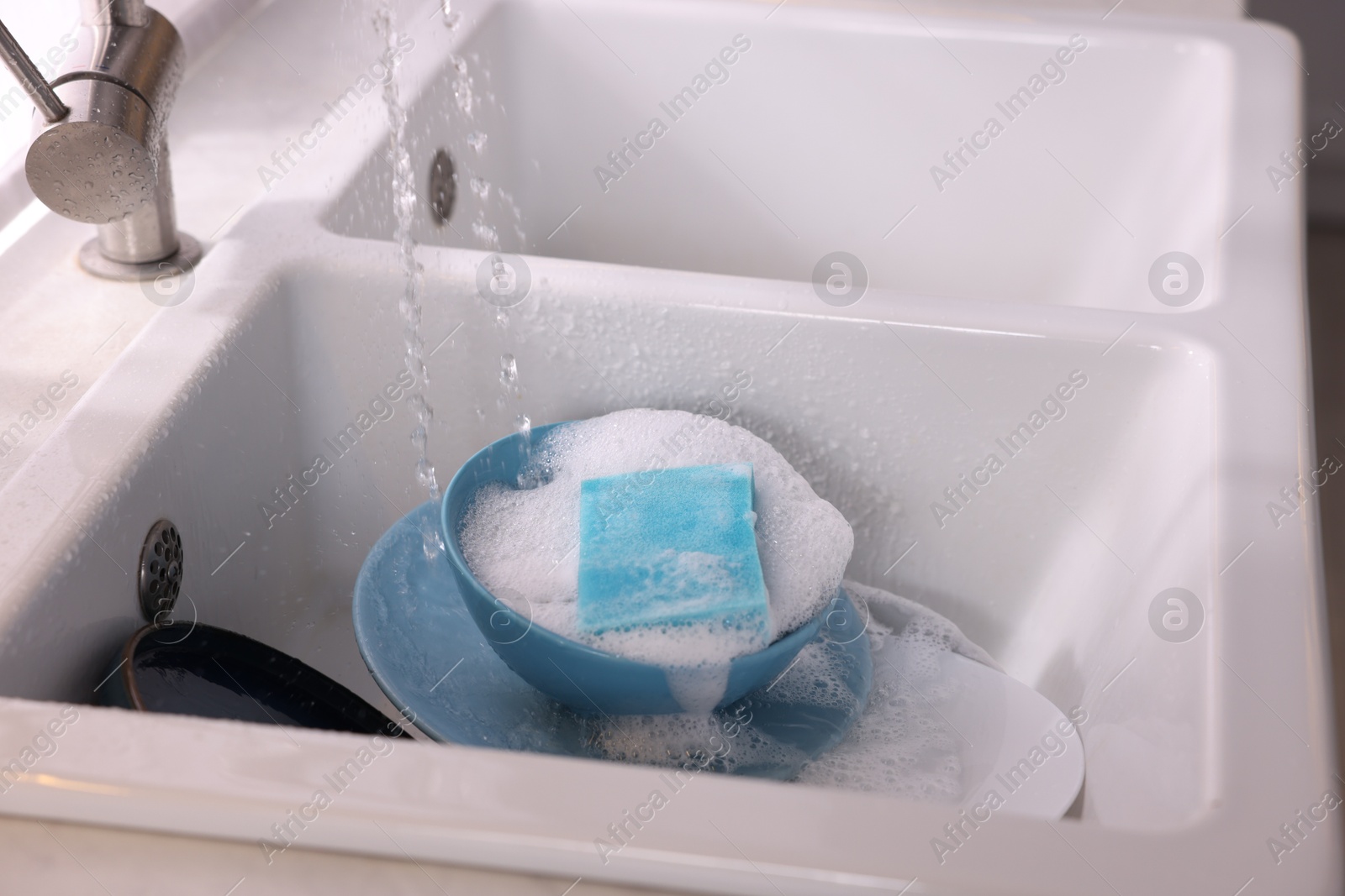 Photo of Dishes, blue sponge and foam under tap water in kitchen sink, closeup