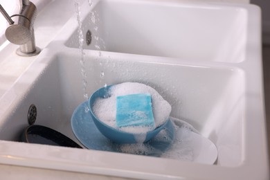 Photo of Dishes, blue sponge and foam under tap water in kitchen sink, closeup