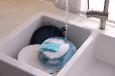 Photo of Dishes, blue sponge and foam under tap water in kitchen sink, closeup