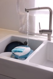 Photo of Dishes, blue sponge and foam under tap water in kitchen sink, closeup