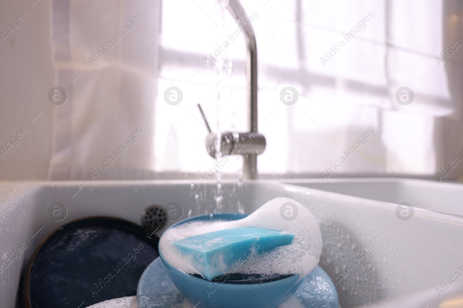 Photo of Dishes, blue sponge and foam under tap water in kitchen sink, closeup