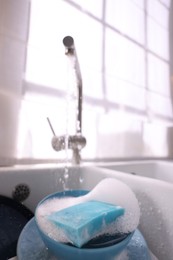 Photo of Dishes, blue sponge and foam under tap water in kitchen sink, closeup