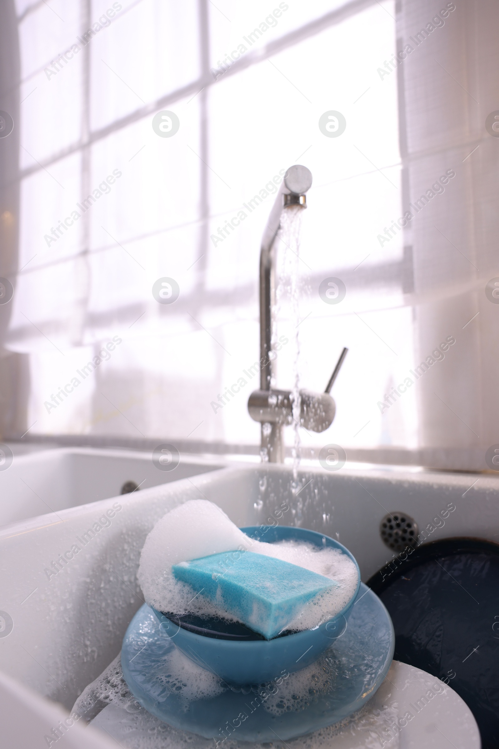 Photo of Dishes, blue sponge and foam under tap water in kitchen sink, closeup