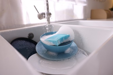 Photo of Dishes, blue sponge and foam under tap water in kitchen sink, closeup