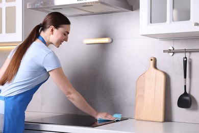 Photo of Smiling woman cleaning induction cooktop with rag in kitchen