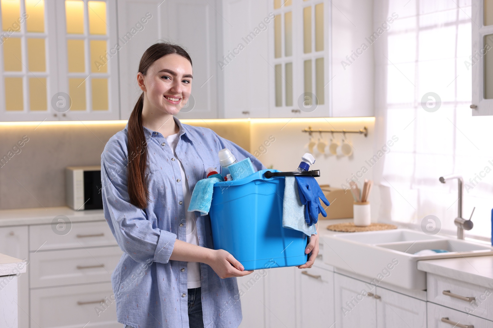 Photo of Smiling woman holding bucket with cleaning supplies in kitchen