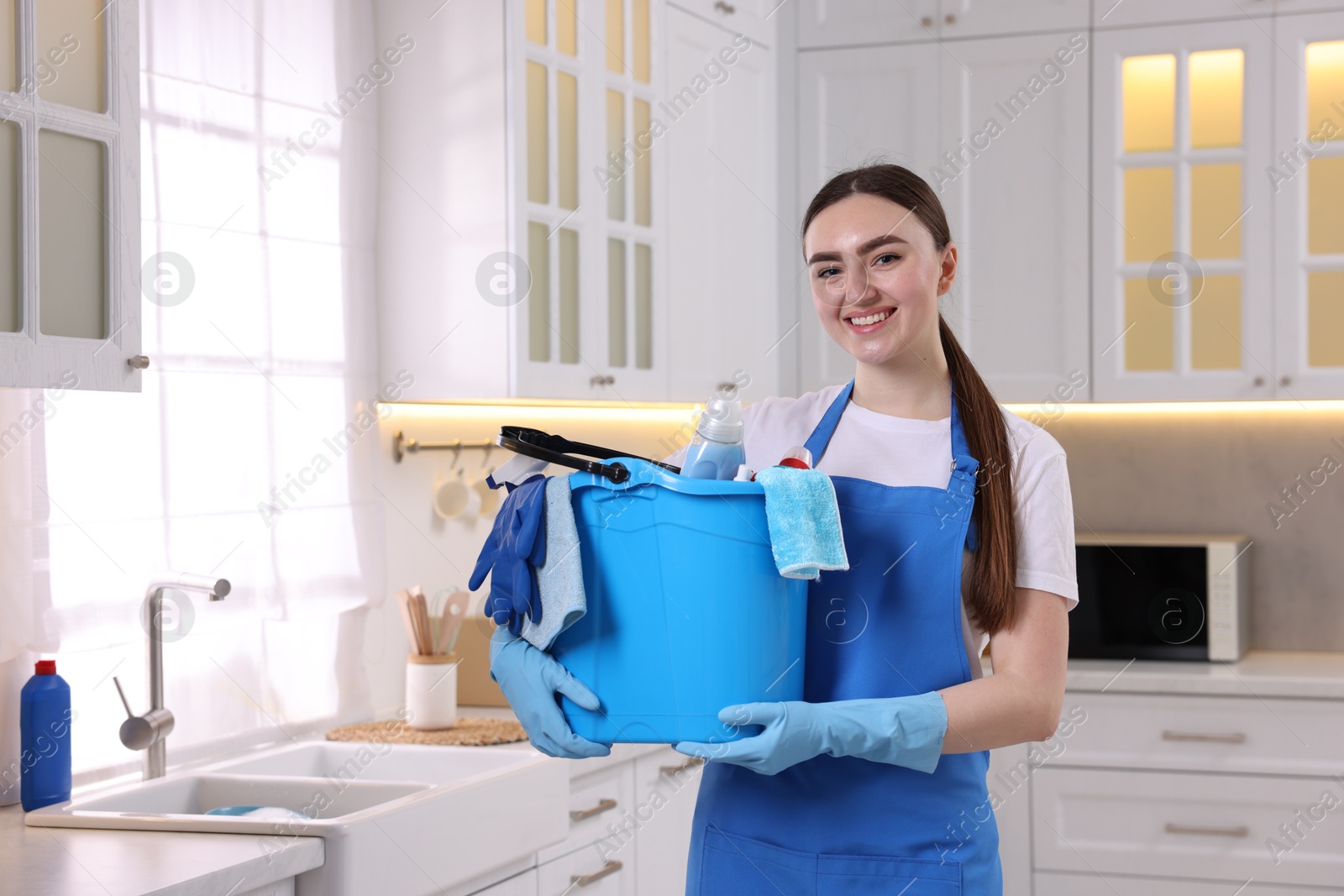 Photo of Smiling woman holding bucket with cleaning supplies in kitchen