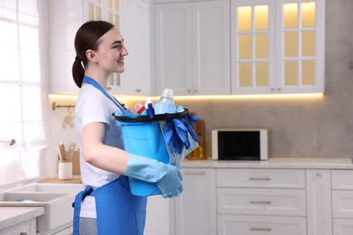 Photo of Smiling woman holding bucket with cleaning supplies in kitchen