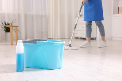 Photo of Woman cleaning floor with string mop indoors, focus on bucket and bottle of detergent