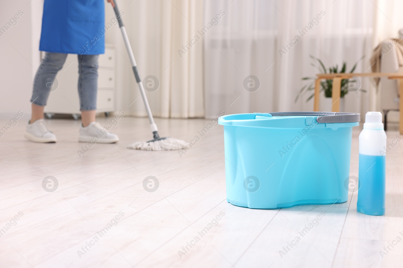 Photo of Woman cleaning floor with string mop indoors, focus on bucket and bottle of detergent