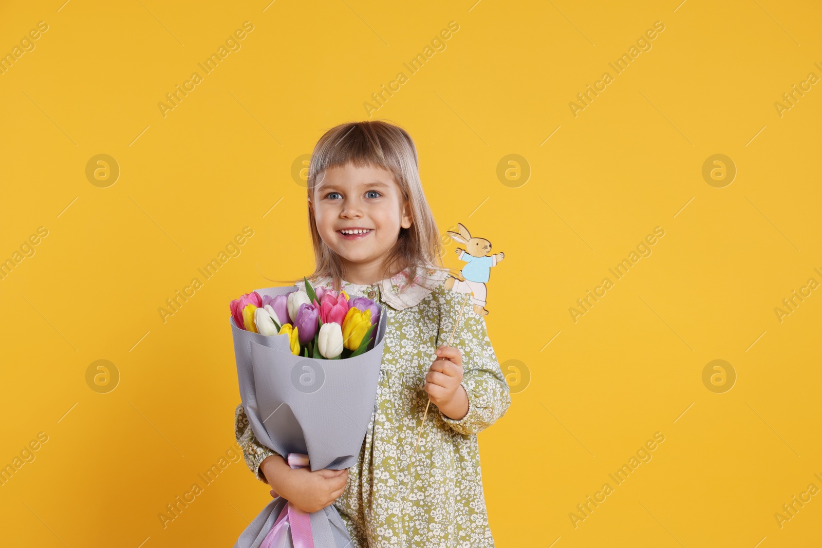 Photo of Smiling little girl with bouquet of tulips and decorative Easter bunny on orange background