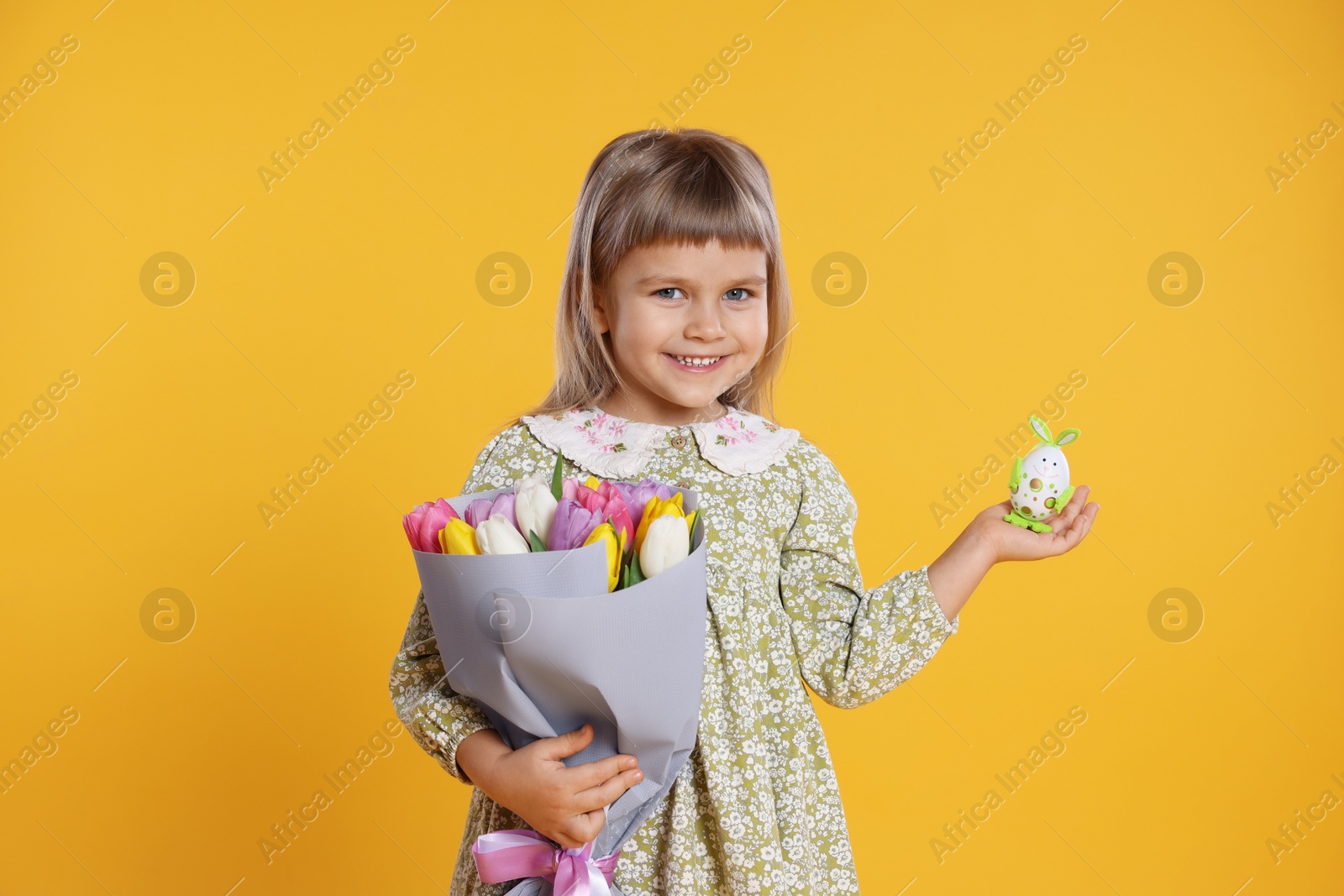 Photo of Smiling little girl with bouquet of tulips and decorative Easter egg on orange background