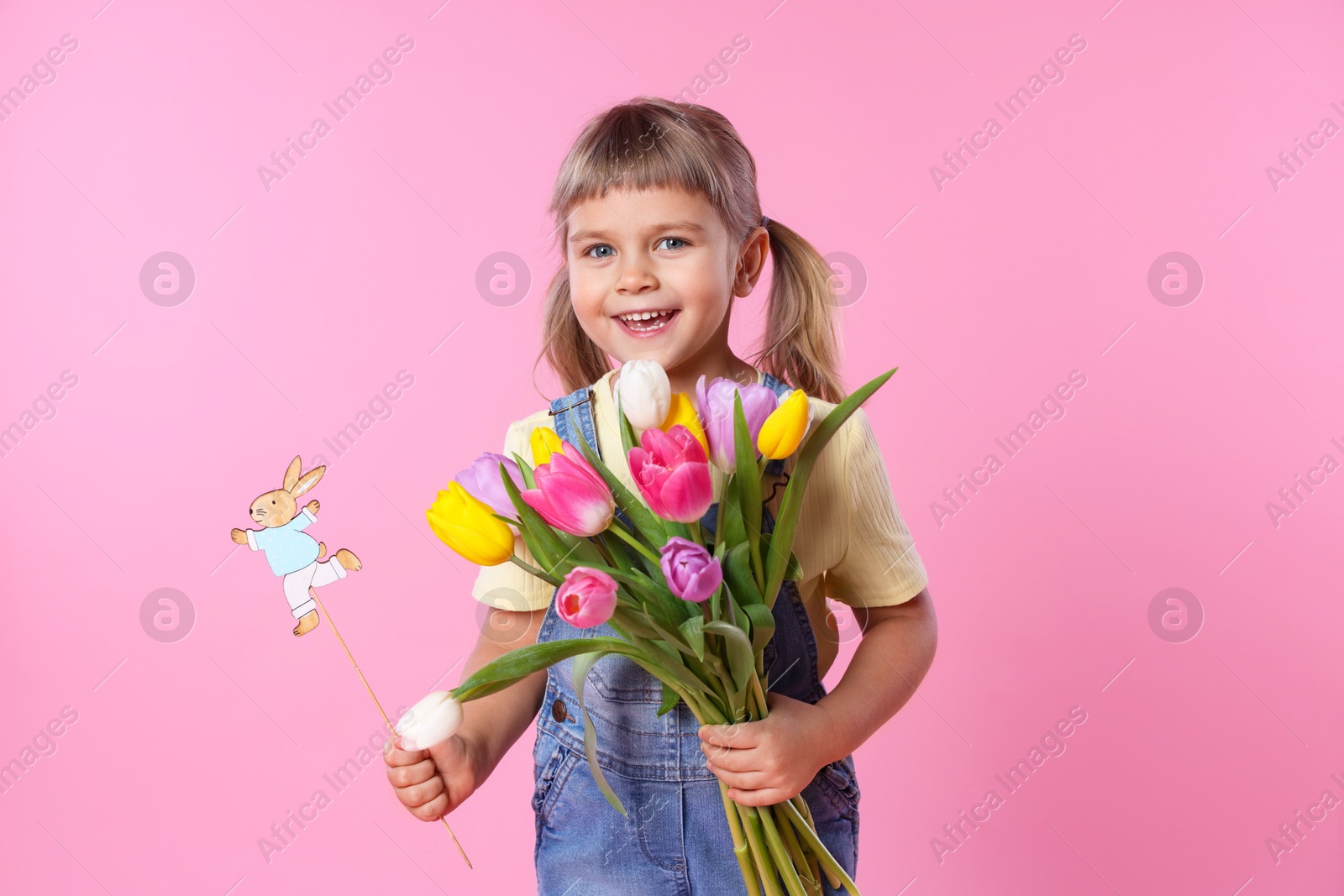 Photo of Smiling little girl with bouquet of tulips and decorative Easter bunny on pink background