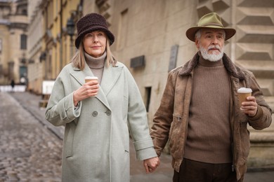 Photo of Lovely senior couple with paper cups holding hands on city street