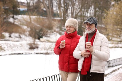 Photo of Happy senior couple with paper cups walking in winter park. Space for text