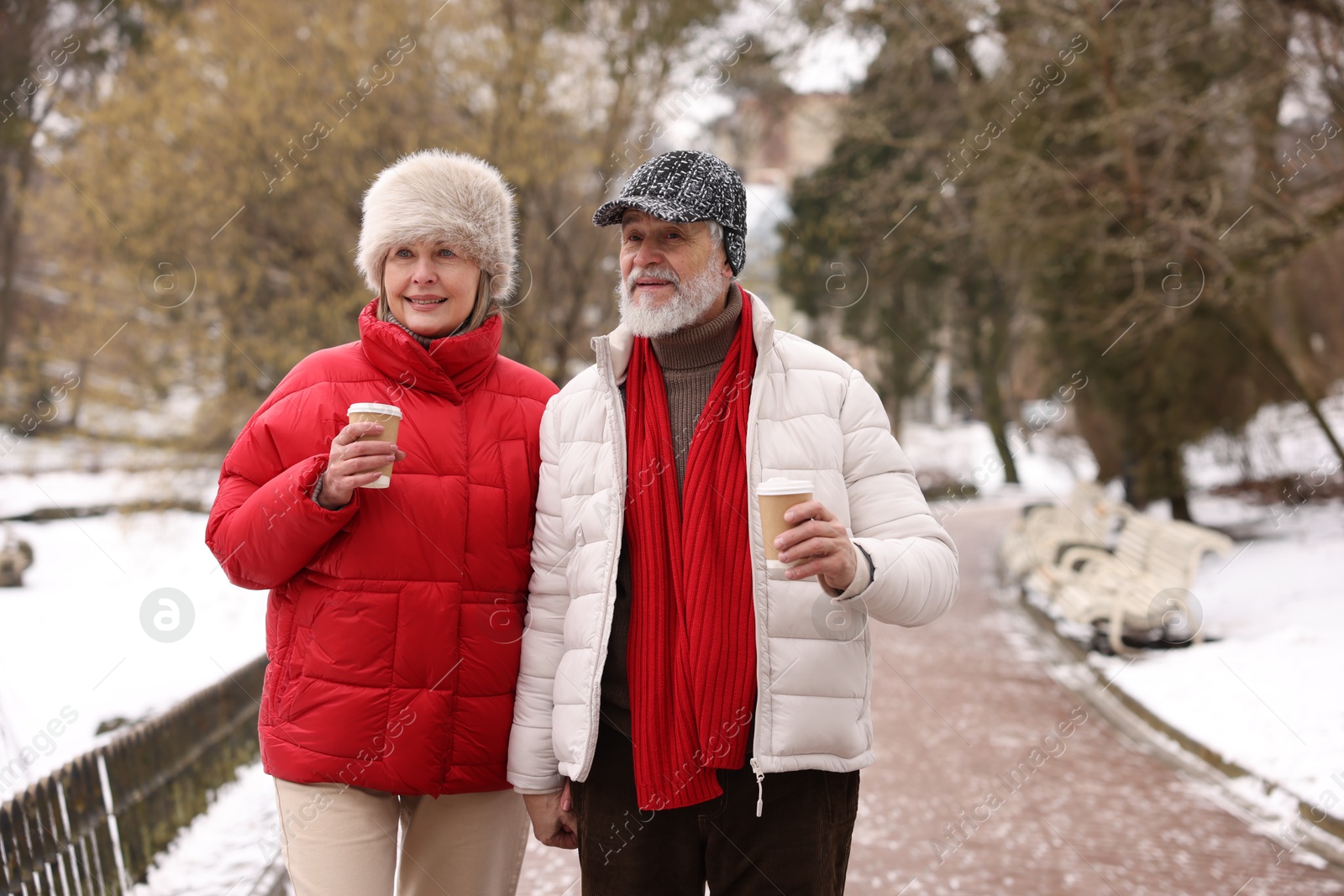 Photo of Happy senior couple with paper cups walking in winter park. Space for text