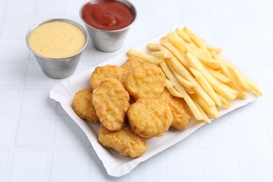 Photo of Tasty chicken nuggets and french fries with sauce on white tiled table, closeup