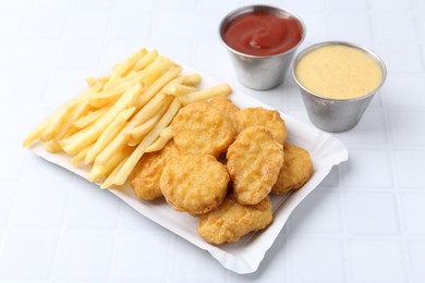 Photo of Tasty chicken nuggets and french fries with sauce on white tiled table, closeup