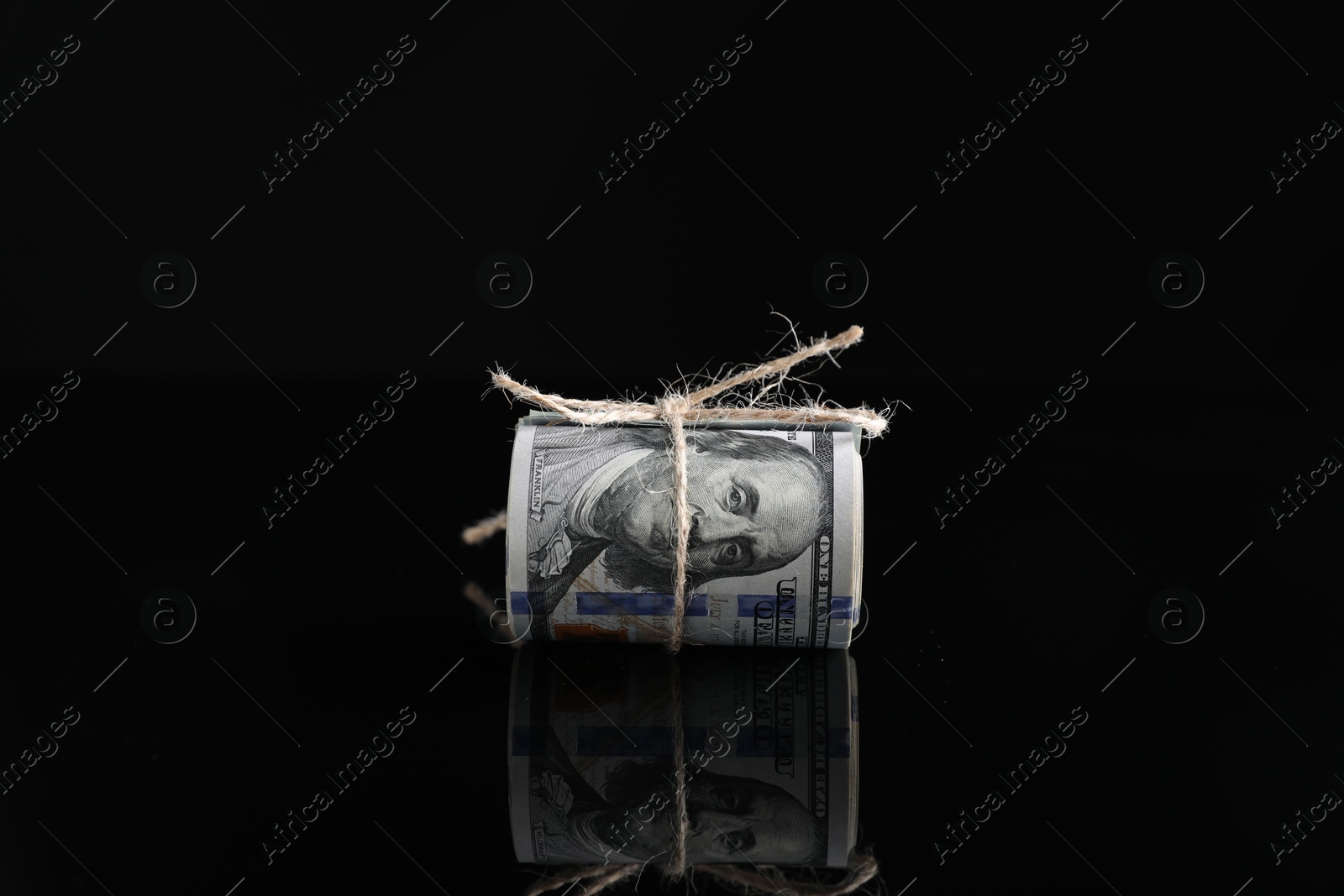 Photo of Rolled dollar banknotes with rope on dark mirror surface