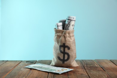 Photo of Burlap sack with dollar banknotes on wooden table against light blue background