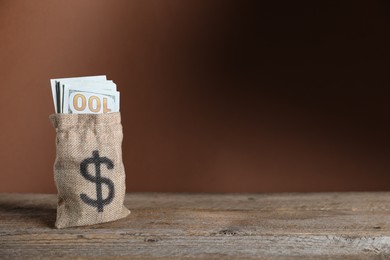 Photo of Dollar banknotes in burlap sack on wooden table against brown background, space for text