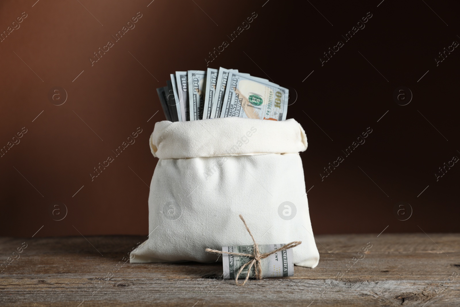 Photo of Burlap sack with dollar banknotes on wooden table against brown background