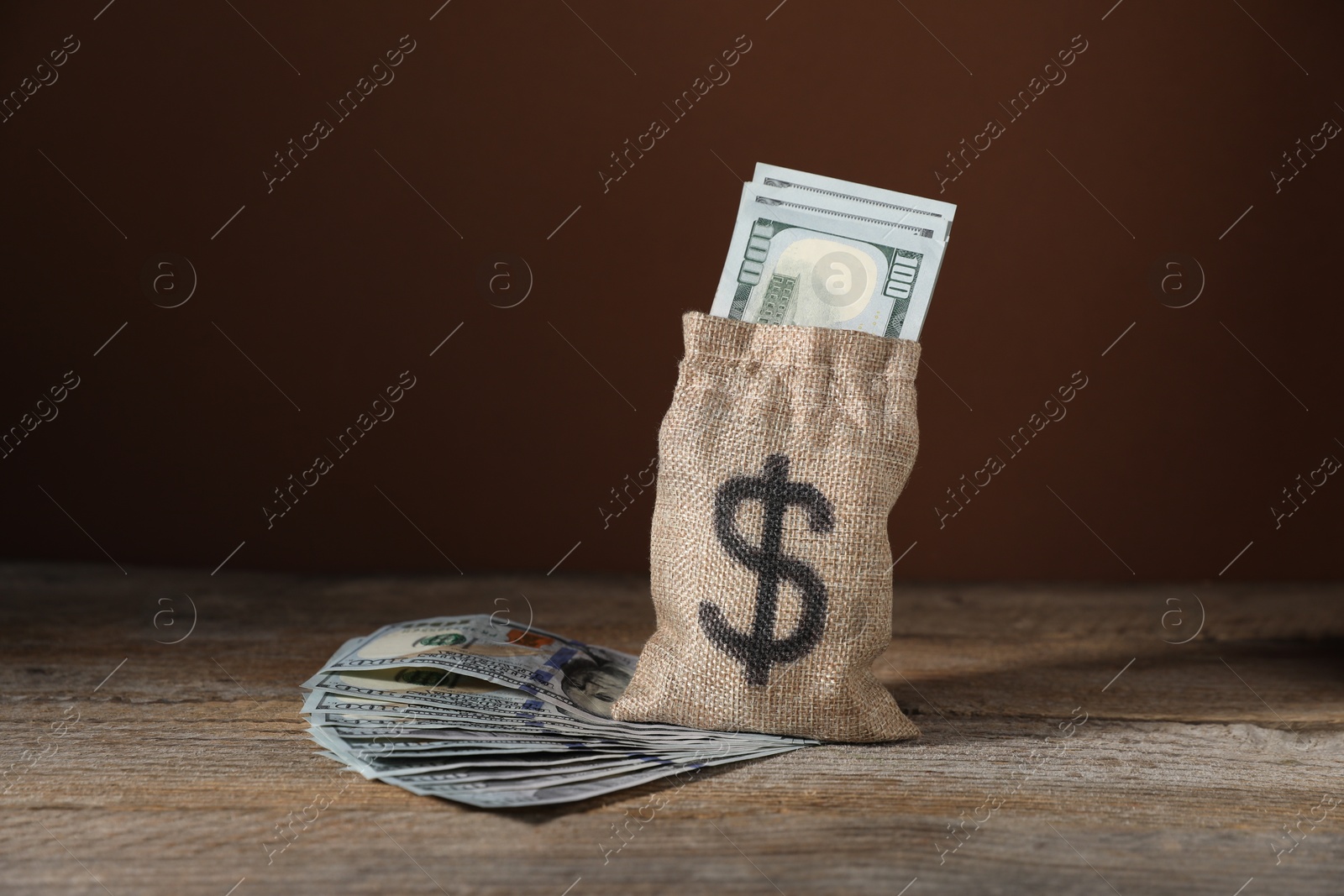 Photo of Burlap sack with dollar banknotes on wooden table against brown background