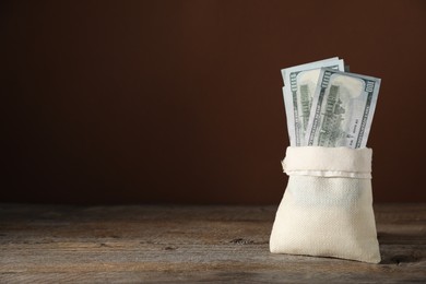 Photo of Dollar banknotes in burlap sack on wooden table against brown background, space for text