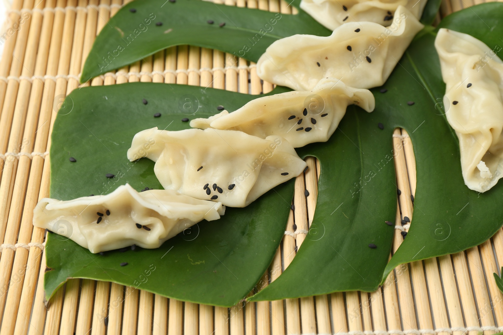 Photo of Tasty boiled gyoza (dumplings) with sesame and monstera leaf on bamboo mat, closeup