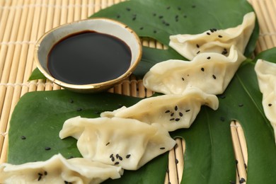 Photo of Tasty boiled gyoza (dumplings) with soy sauce and monstera leaf on bamboo mat, closeup