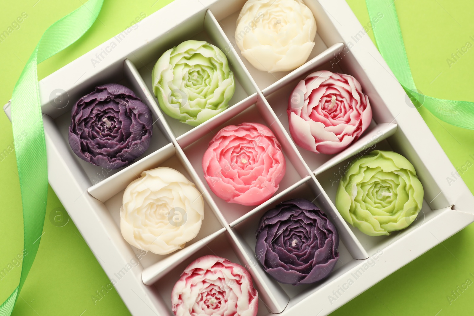 Photo of Beautiful flower-shaped candles in box and ribbon on light green background, top view