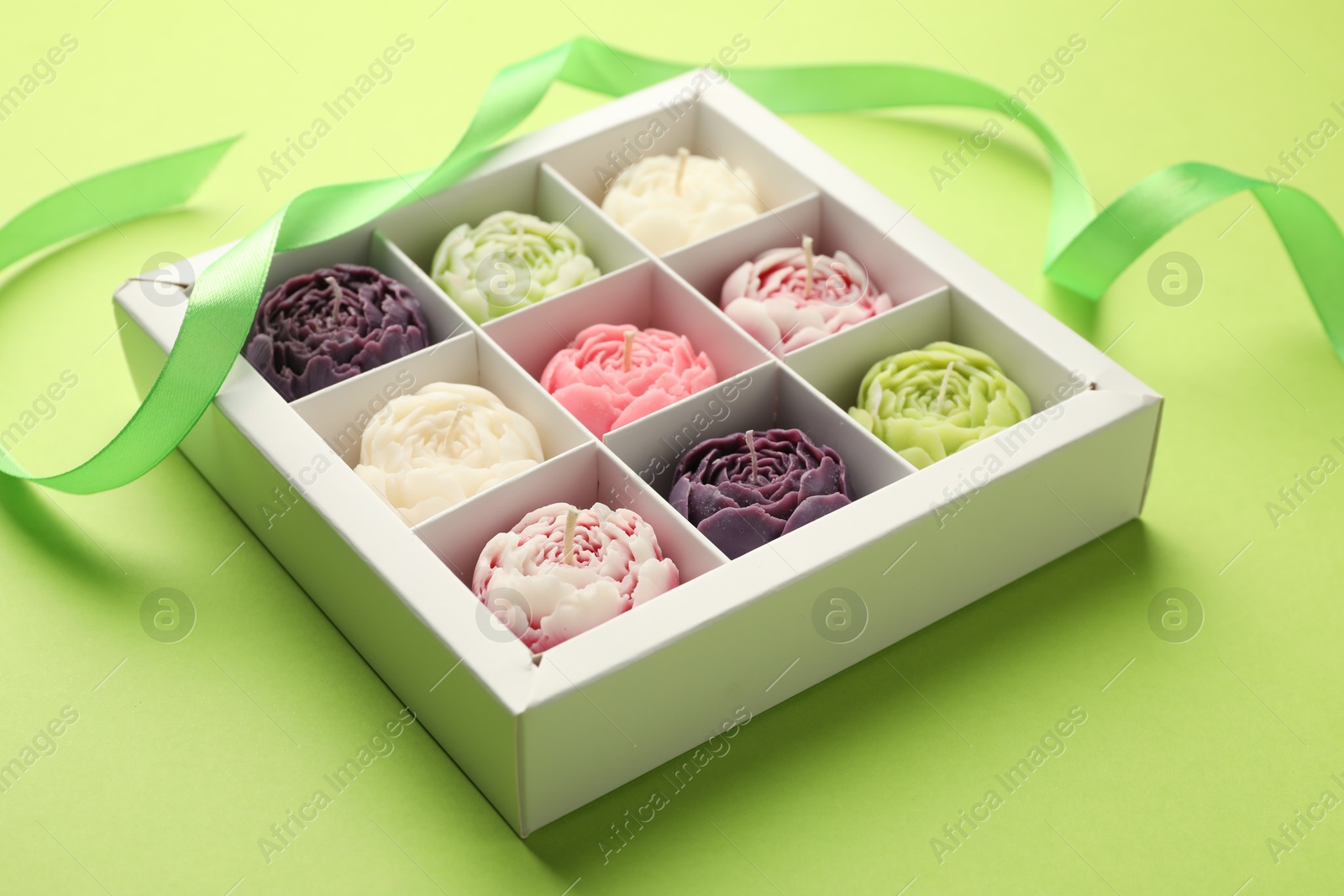 Photo of Beautiful flower-shaped candles in box and ribbon on light green background, closeup