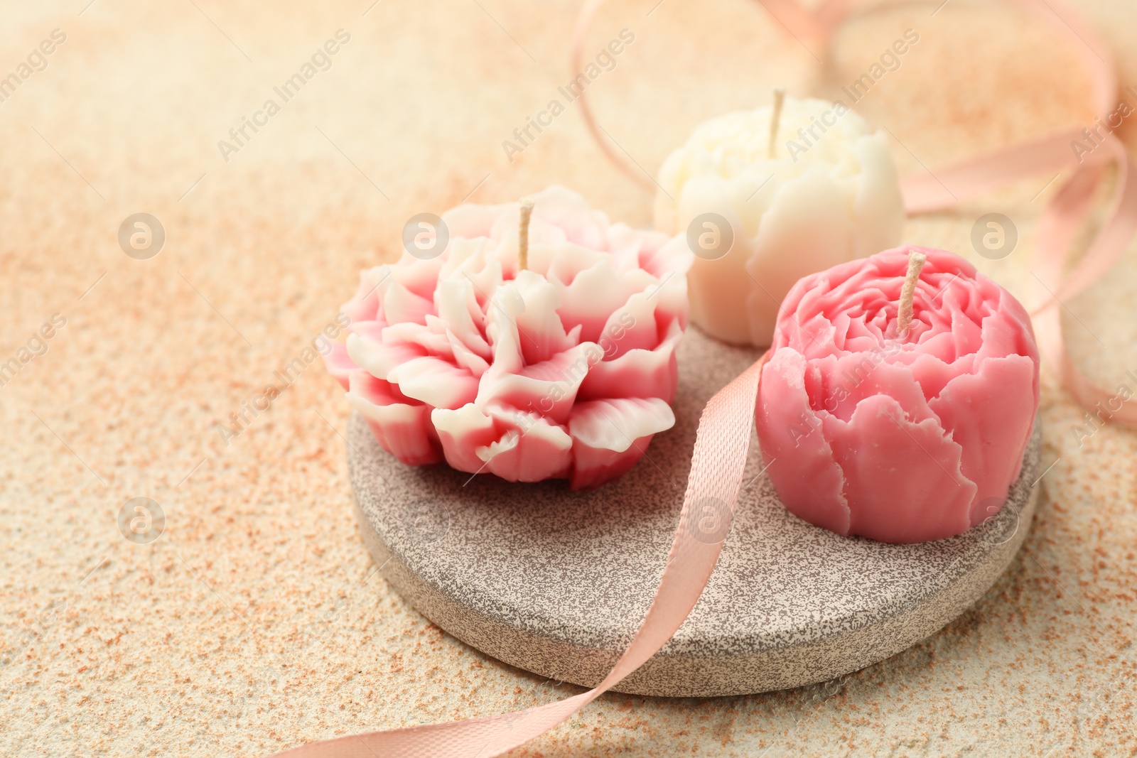 Photo of Beautiful flower-shaped candles and ribbon on beige textured background, closeup