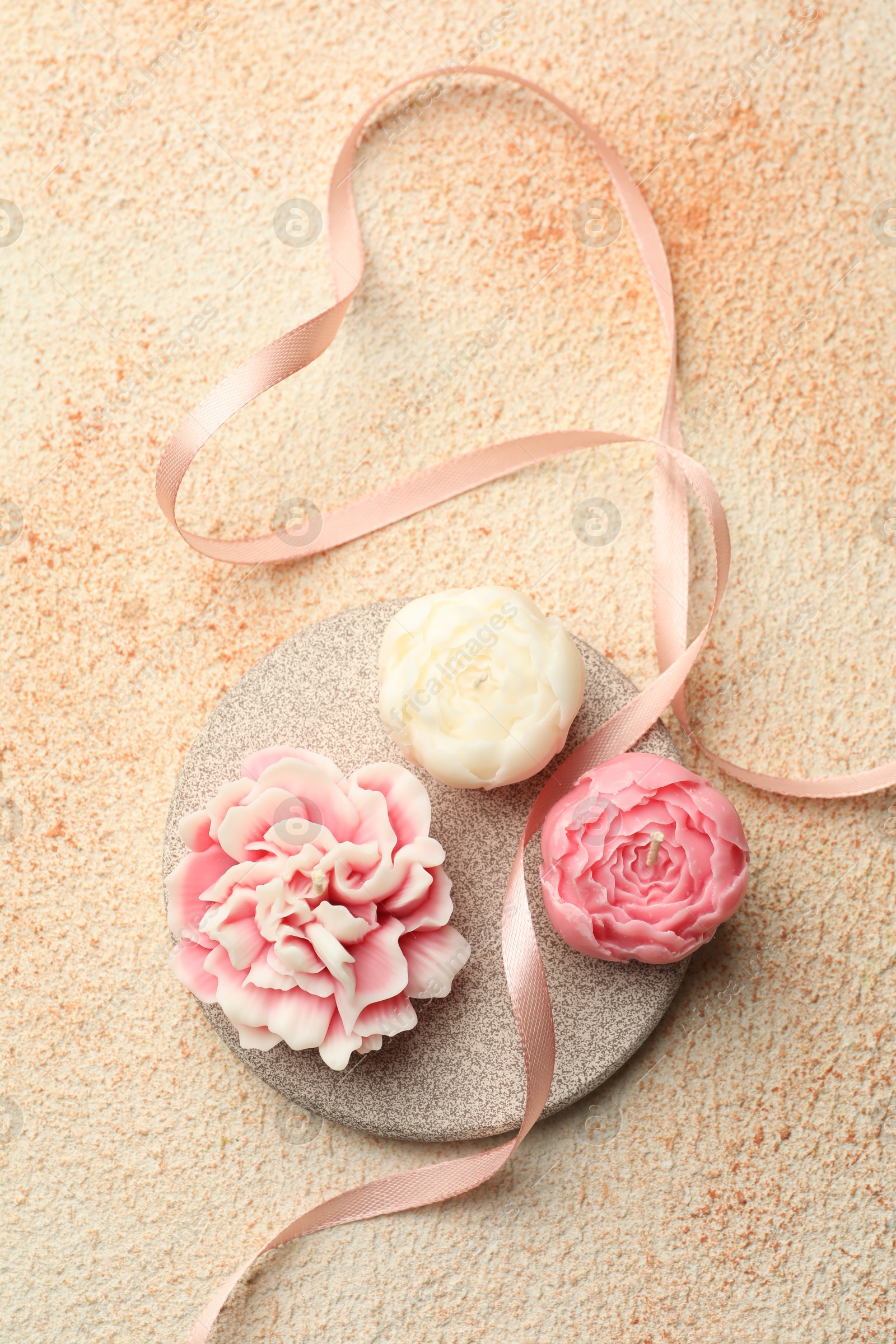 Photo of Beautiful flower-shaped candles and ribbon on beige textured background, top view