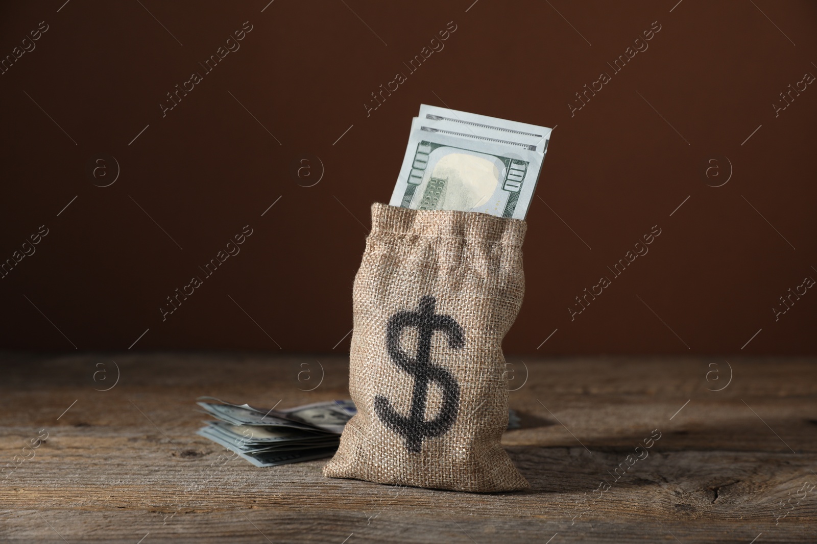 Photo of Burlap sack with dollar banknotes on wooden table against brown background