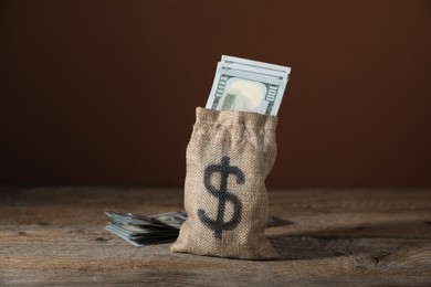 Photo of Burlap sack with dollar banknotes on wooden table against brown background