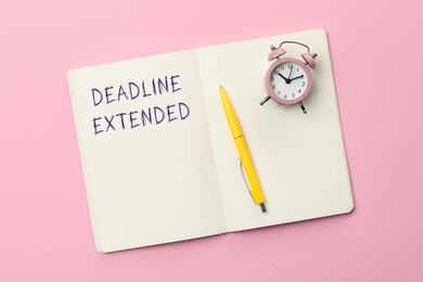 Image of Text Deadline Extended in notebook, pen and alarm clock on pink background, top view