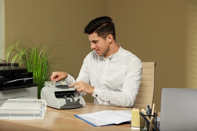 Photo of Man putting money into banknote counter at wooden table indoors