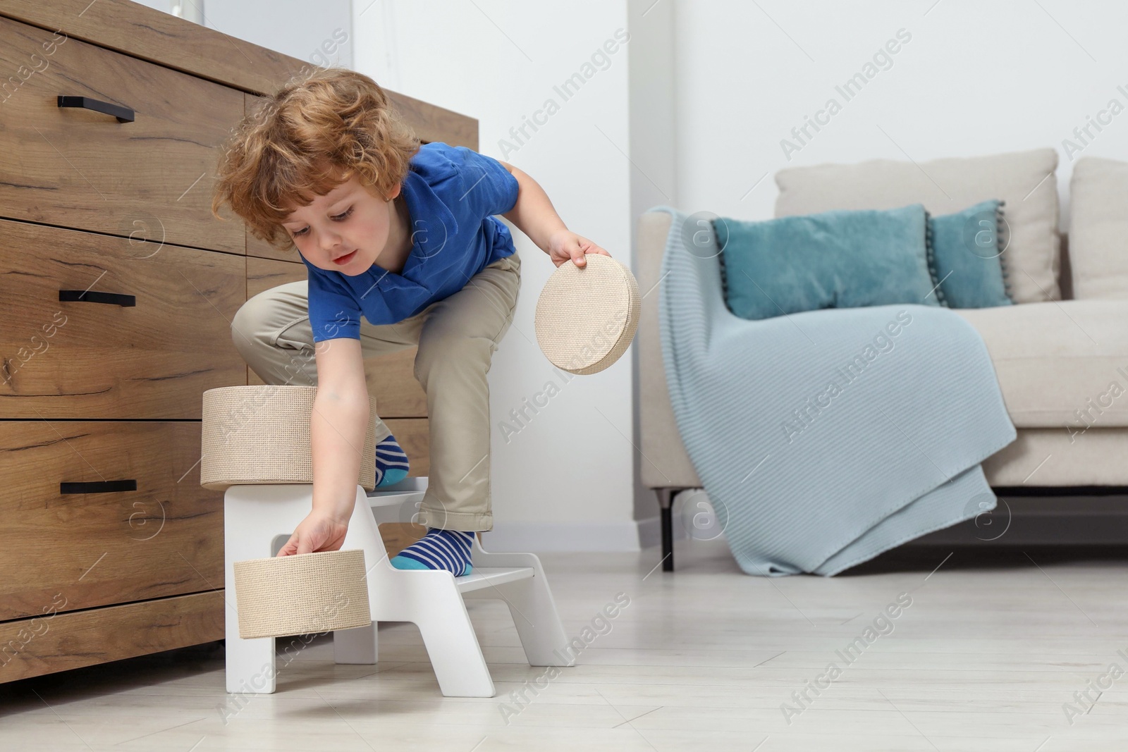 Photo of Little boy with box standing on step stool near chest of drawers at home