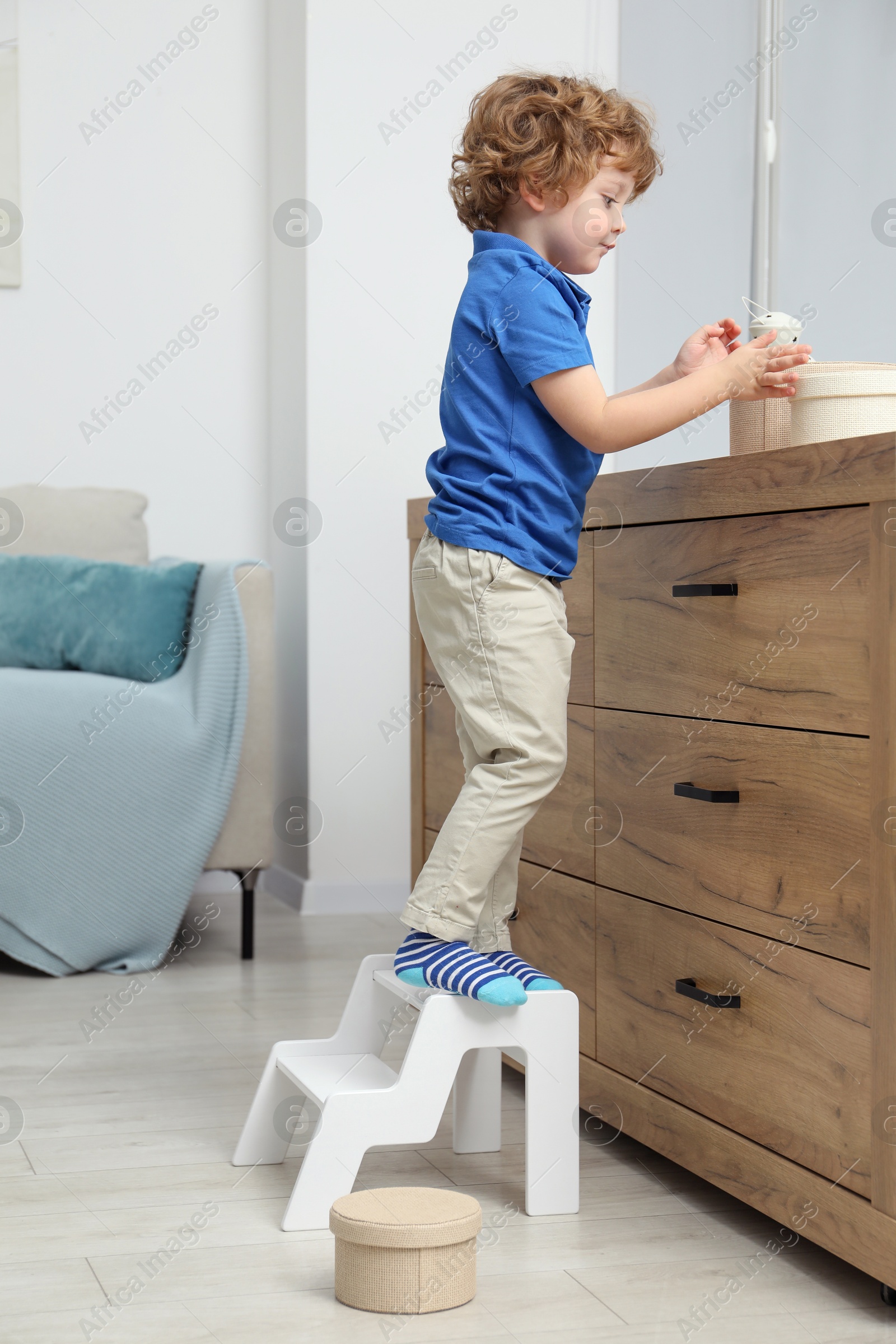 Photo of Little boy standing on step stool and reaching for box at home