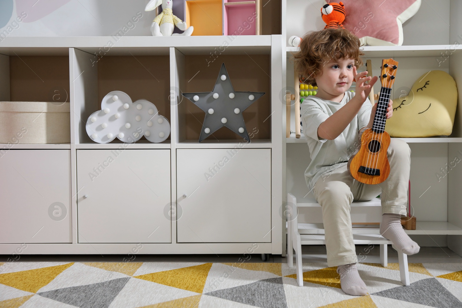 Photo of Little boy with toy guitar sitting on step stool at home