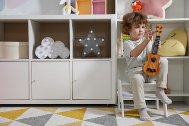 Photo of Little boy with toy guitar sitting on step stool at home