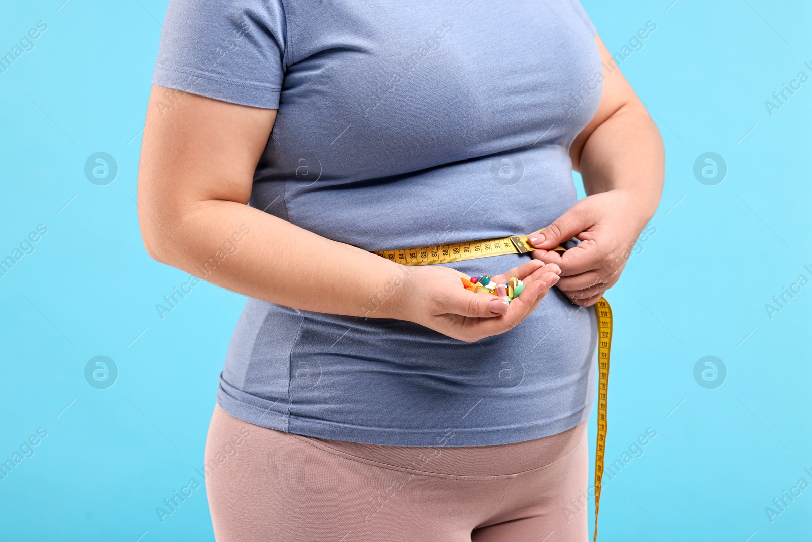 Photo of Plus size woman measuring waist with tape and holding pile of weight loss supplements on light blue background, closeup