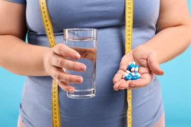 Photo of Plus size woman holding pile of weight loss supplements and glass with water on light blue background, closeup