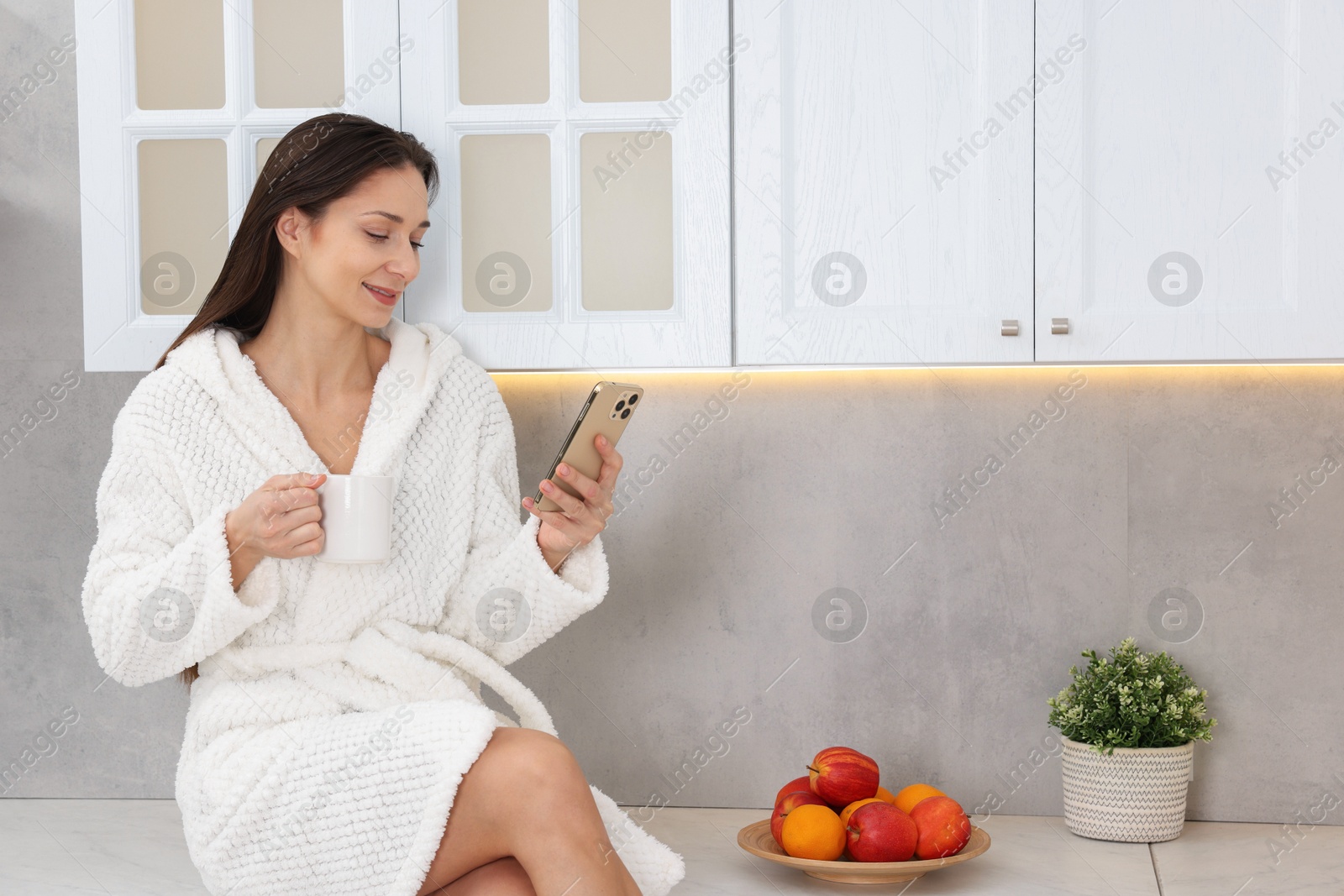 Photo of Beautiful woman with cup of drink using smartphone after spa procedure on countertop in kitchen