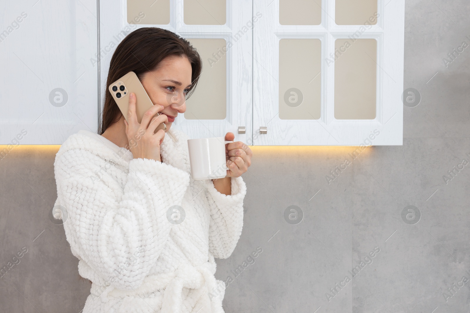 Photo of Beautiful woman with cup of drink talking on smartphone after spa procedure in kitchen