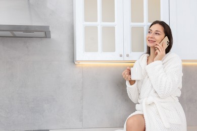 Photo of Beautiful woman with cup of drink talking on smartphone after spa procedure in kitchen