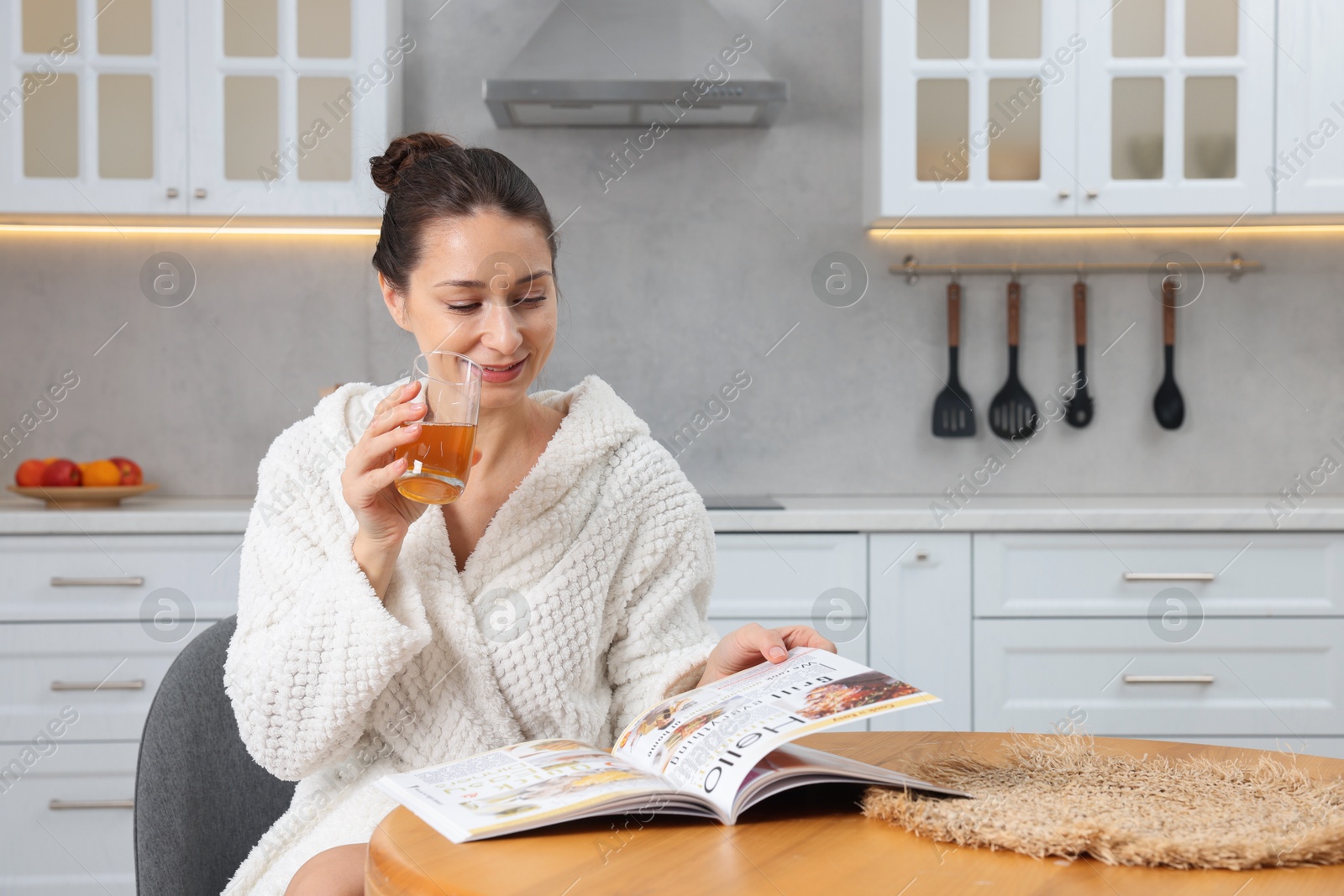 Photo of Beautiful woman with glass of juice reading magazine after spa procedure in kitchen