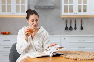 Photo of Beautiful woman with glass of juice reading magazine after spa procedure in kitchen