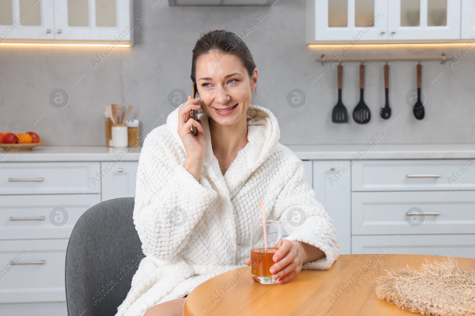 Photo of Beautiful woman with glass of juice talking on smartphone after spa procedure in kitchen